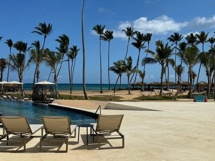 A swimming pool surrounded by chairs and palm trees with the ocean in the background