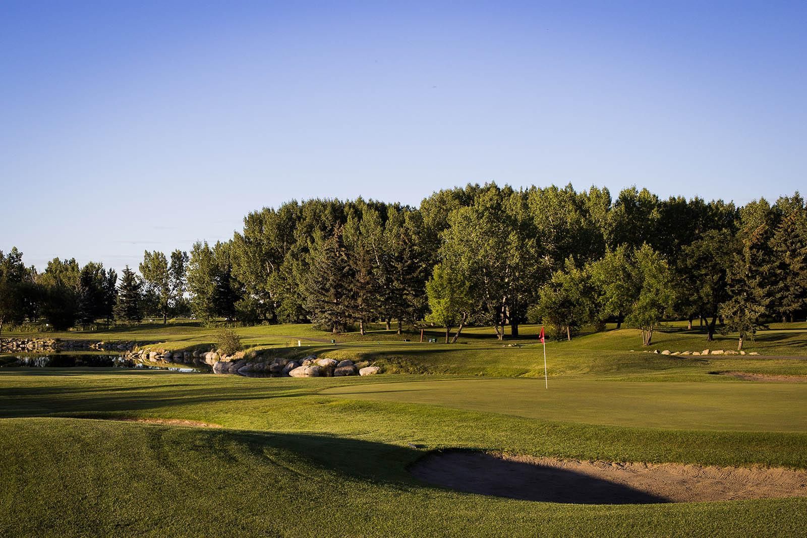 High-angle view of a golf green surrounded by sand traps, with a river and autumn-colored trees in the background.