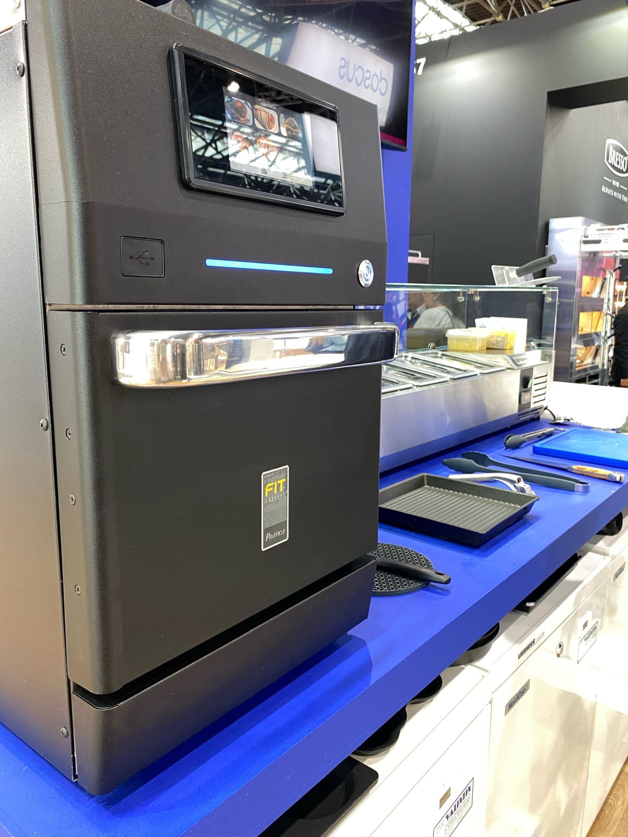 Black commercial oven on a blue counter with a display screen, at a trade show.