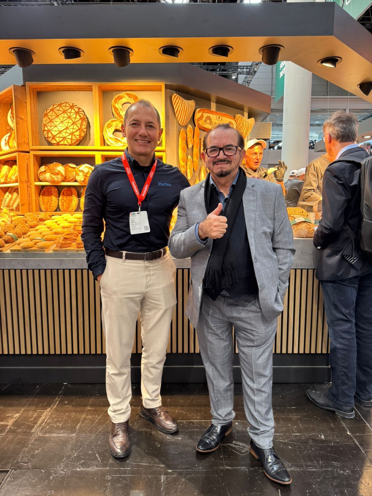 Two men pose in front of a bakery display. One gives a thumbs up, both smile. Wooden shelves with bread behind them.