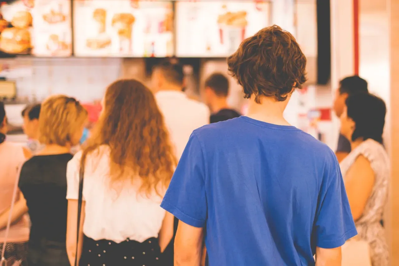 People wait in line at a fast-food restaurant; a young man in a blue shirt is in the foreground.