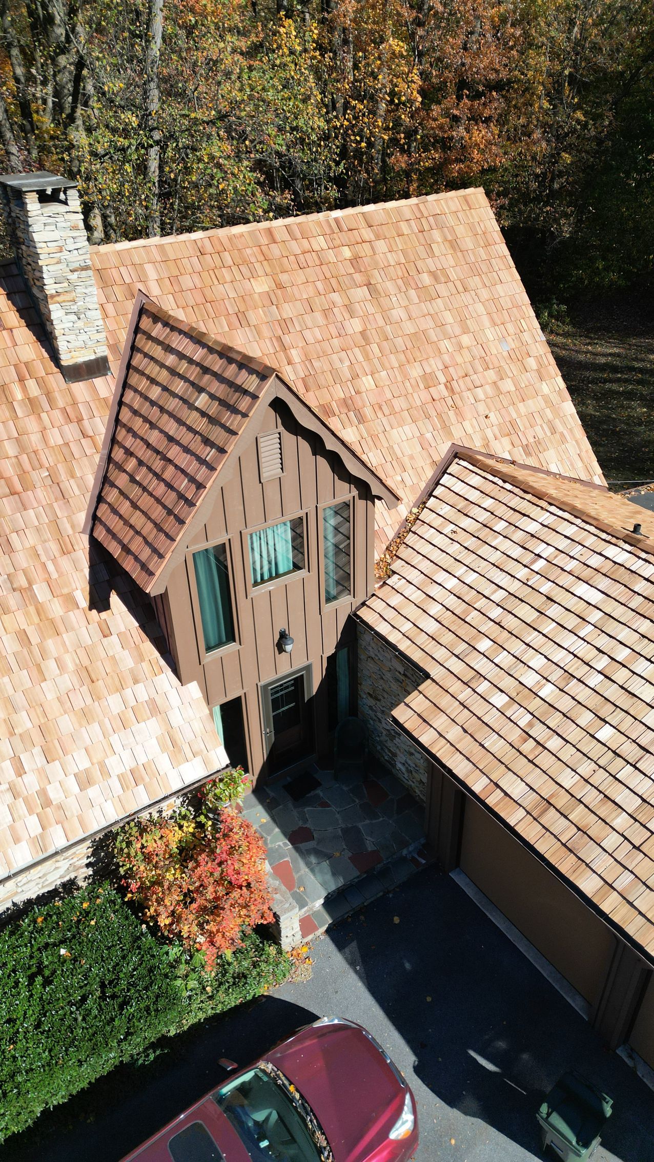Overhead view of a brown shingle-roofed house with a red car parked in the driveway, fall foliage.