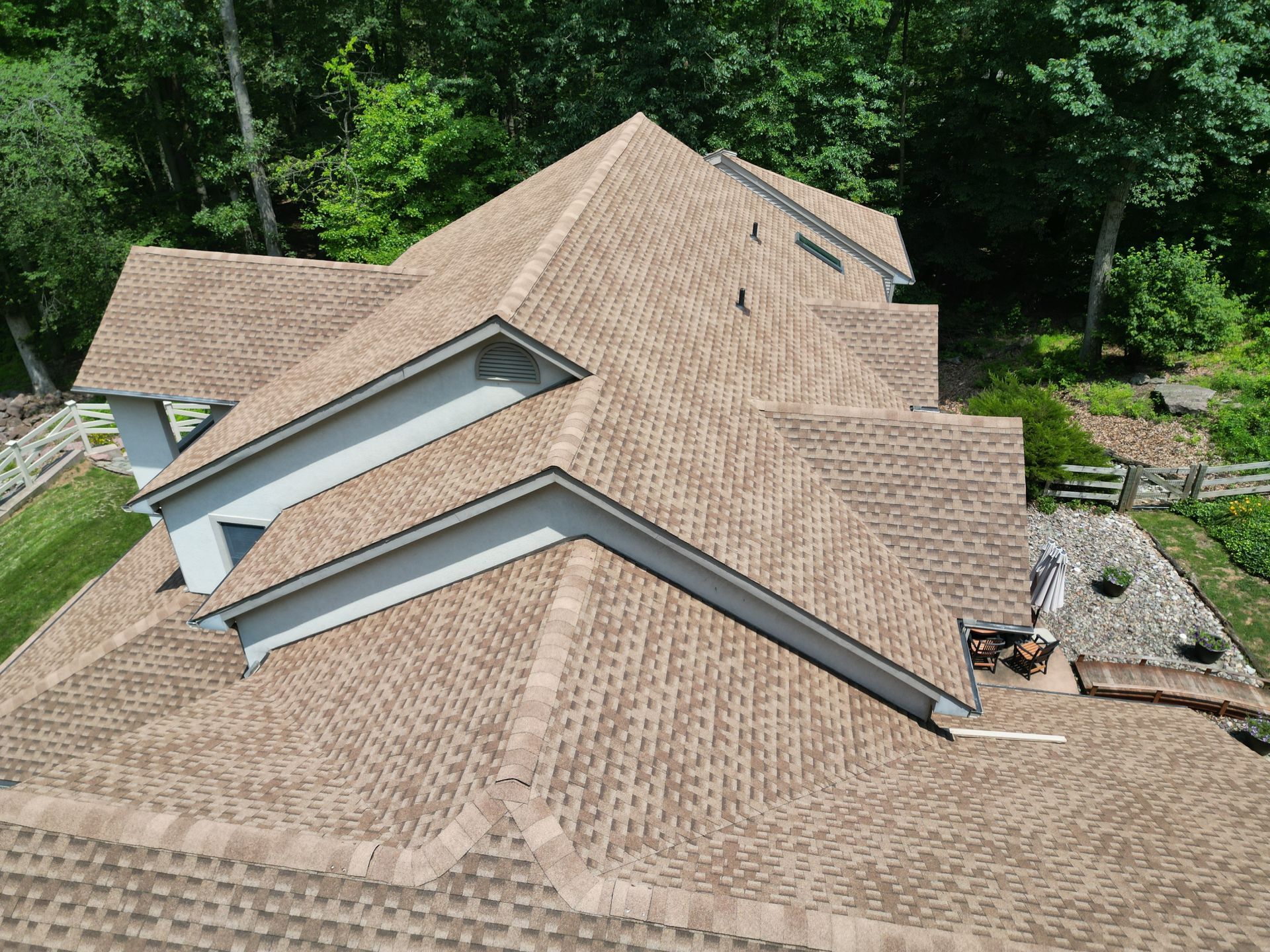 Tan shingle roof on a house, viewed from above, surrounded by trees and greenery.