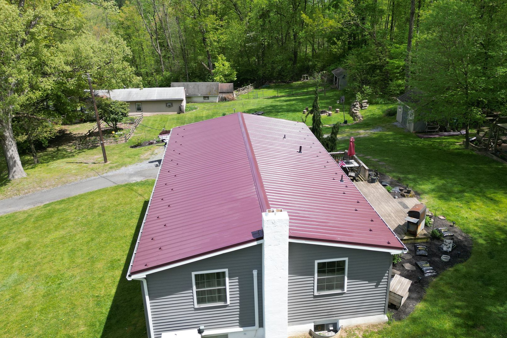 Gray house with a maroon metal roof, chimney, windows, and a deck. Green grass and trees surround the house.