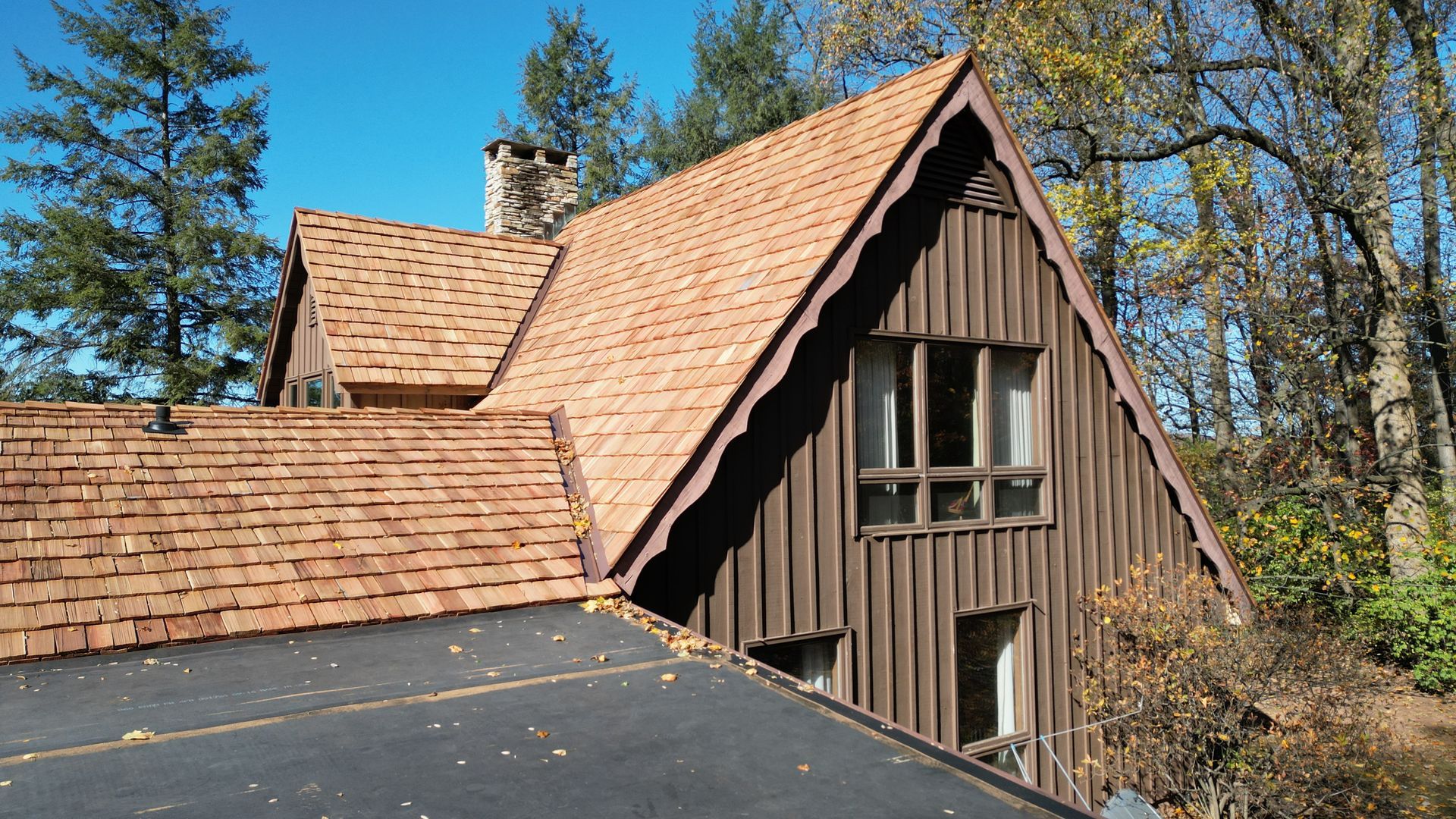 Brown A-frame house with cedar shake roof, set in a wooded area on a sunny day.