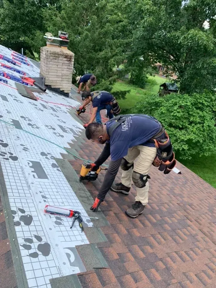 Three roofers installing shingles on a roof. One uses a nail gun, others lay shingles.