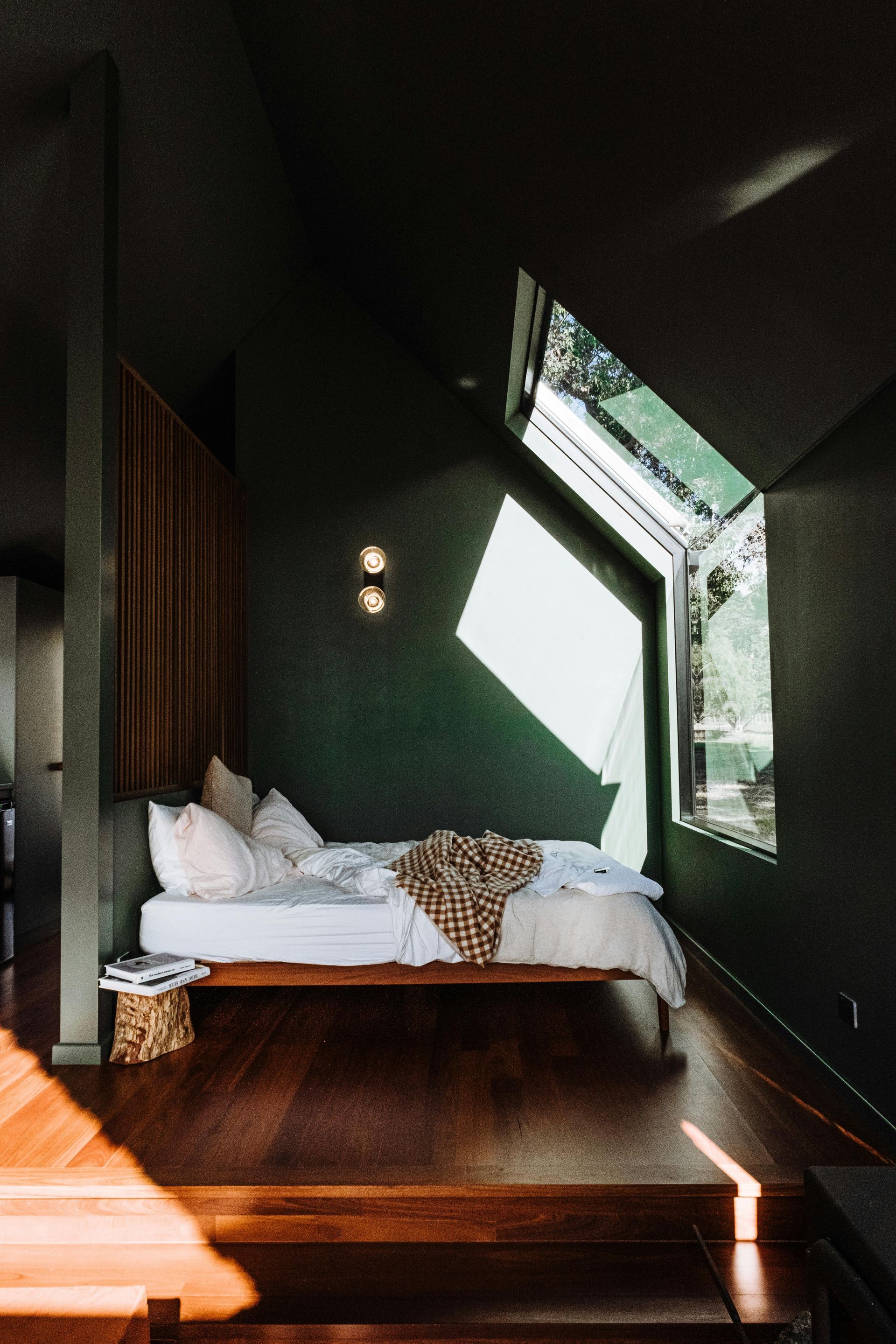 Bedroom with dark green walls, wooden floor, bed, and angular window; sunlight streams in.