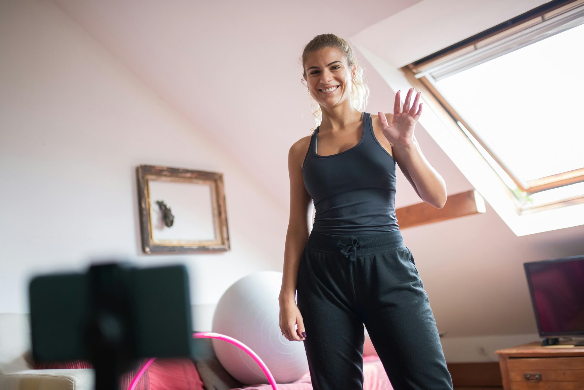 Woman in workout clothes waves at a smartphone, smiling. Indoors with a light-filled window.