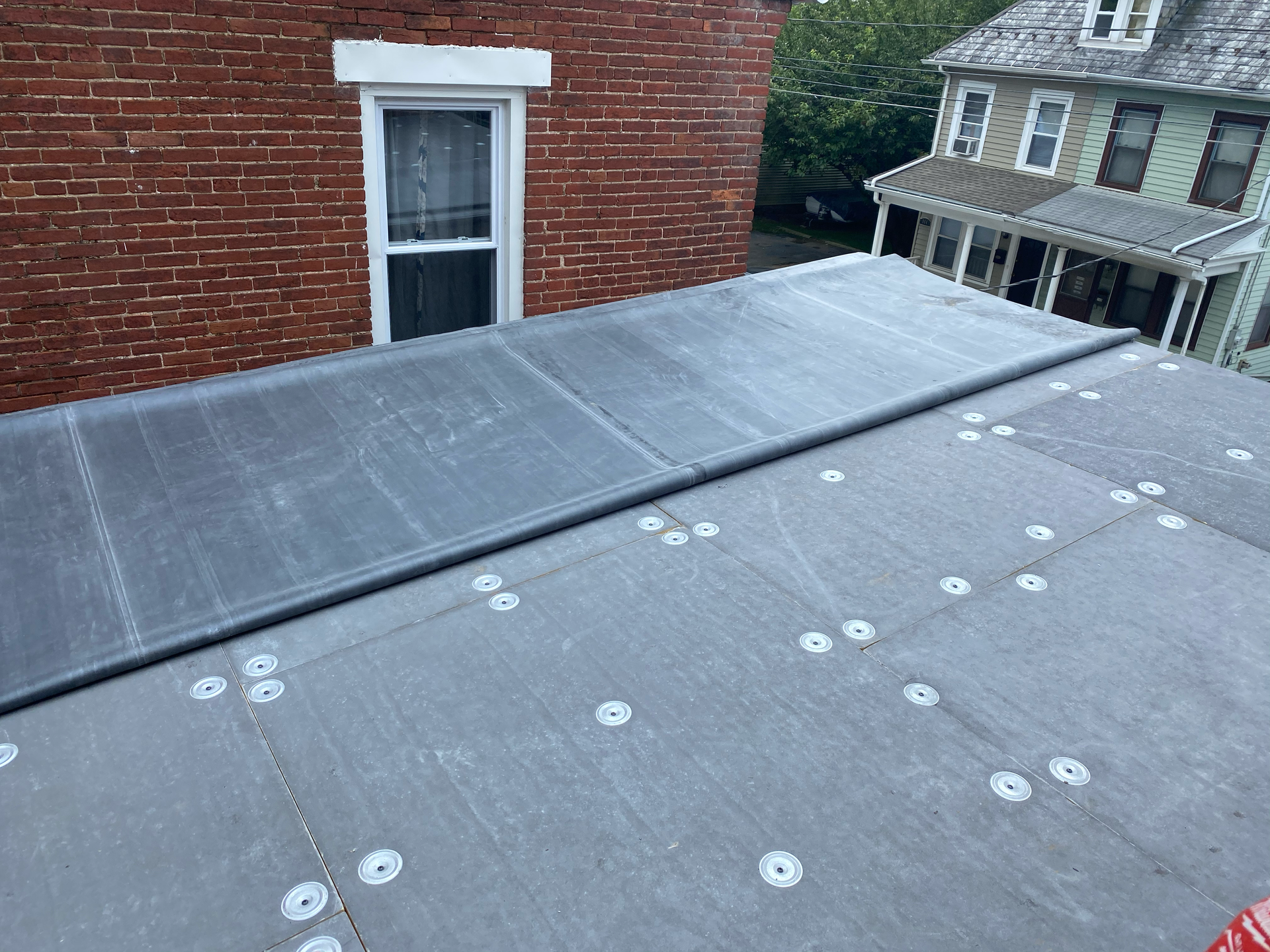 Flat roof with gray material, fasteners, and a building with a brick wall.