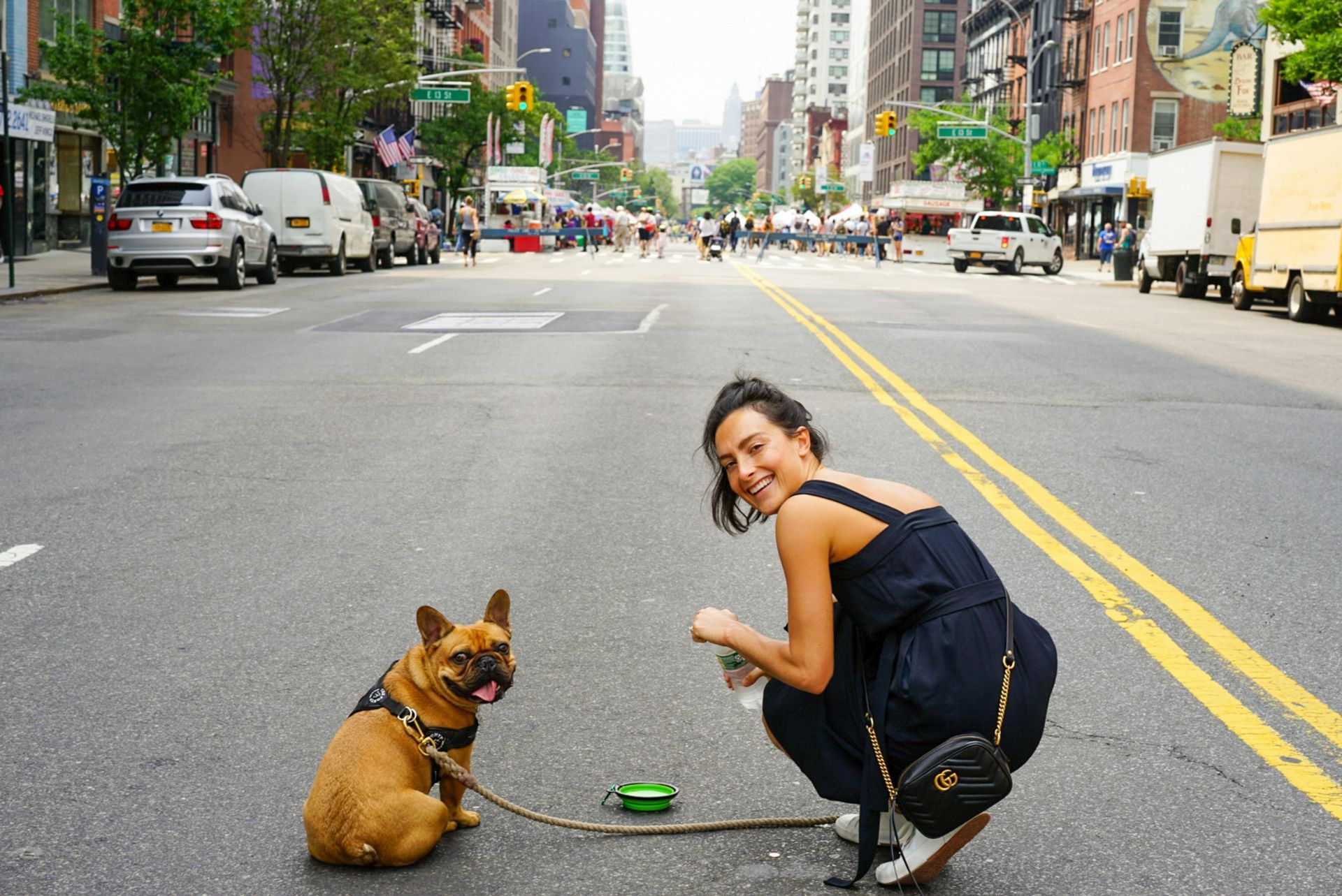 Woman crouches in a city street beside a small brown dog on a leash.