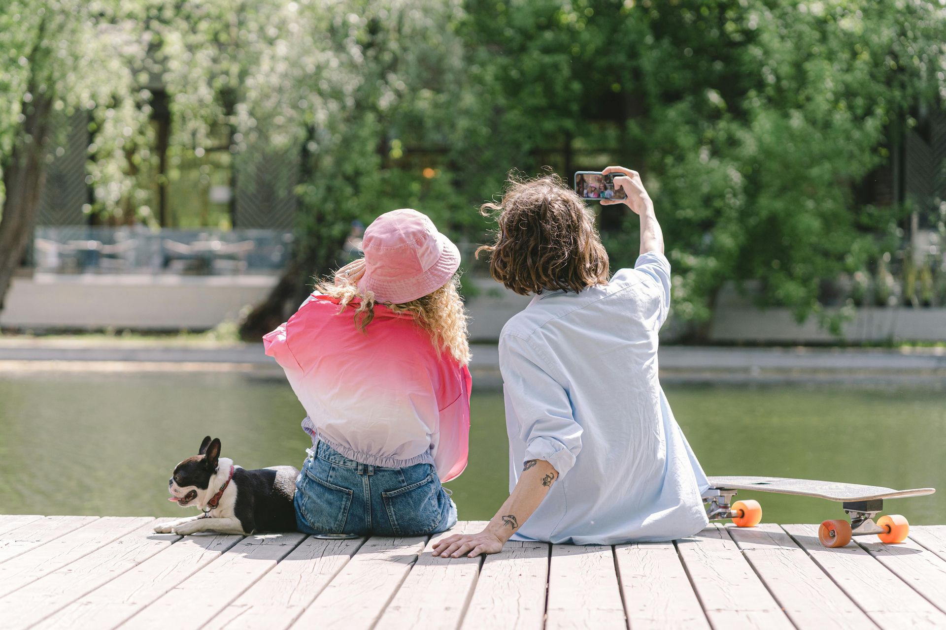 Two people sit on a dock by a lake, one taking a selfie, skateboards beside them.