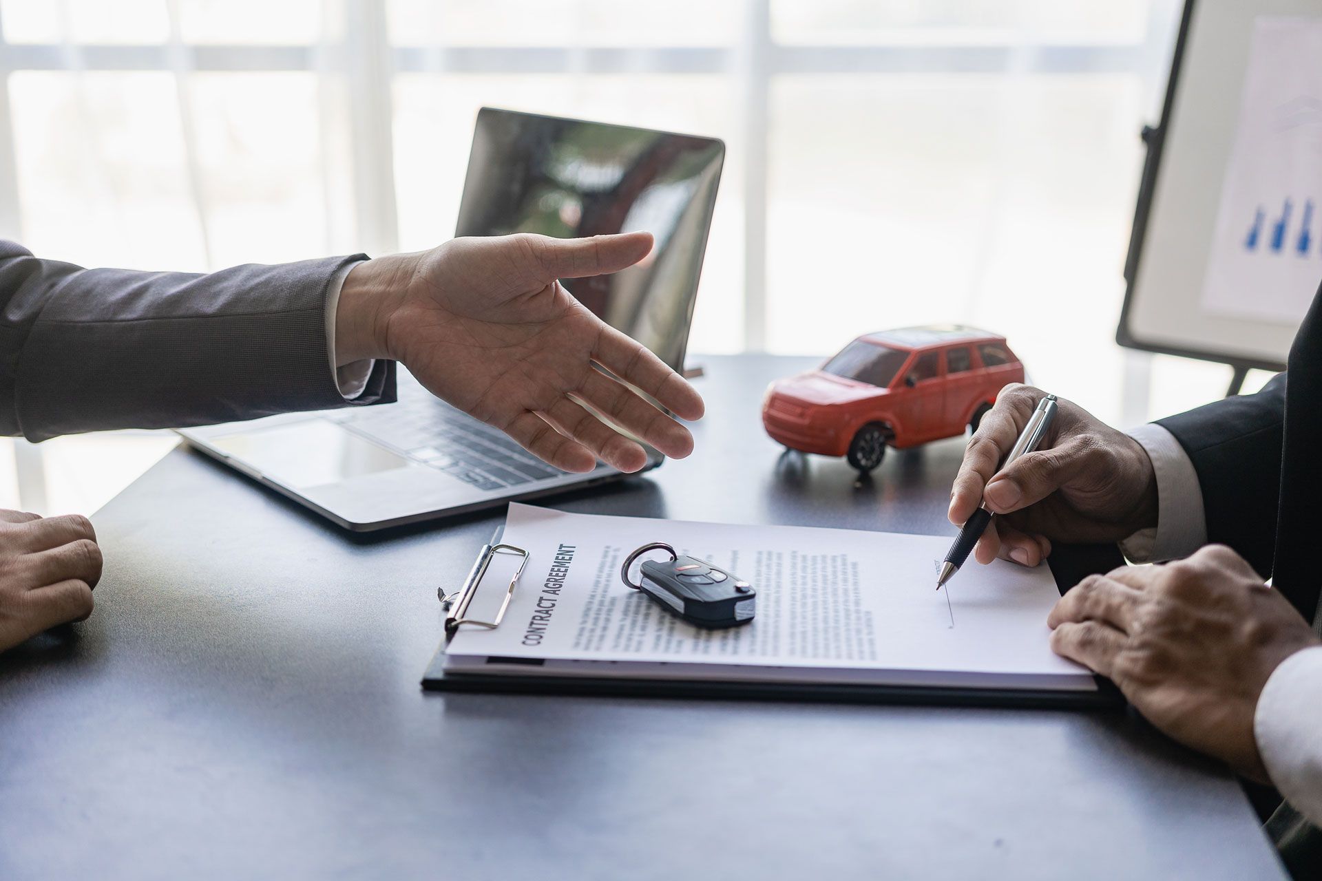 Man signing car purchase agreement, shaking hands with another person at a desk with a laptop and toy car.