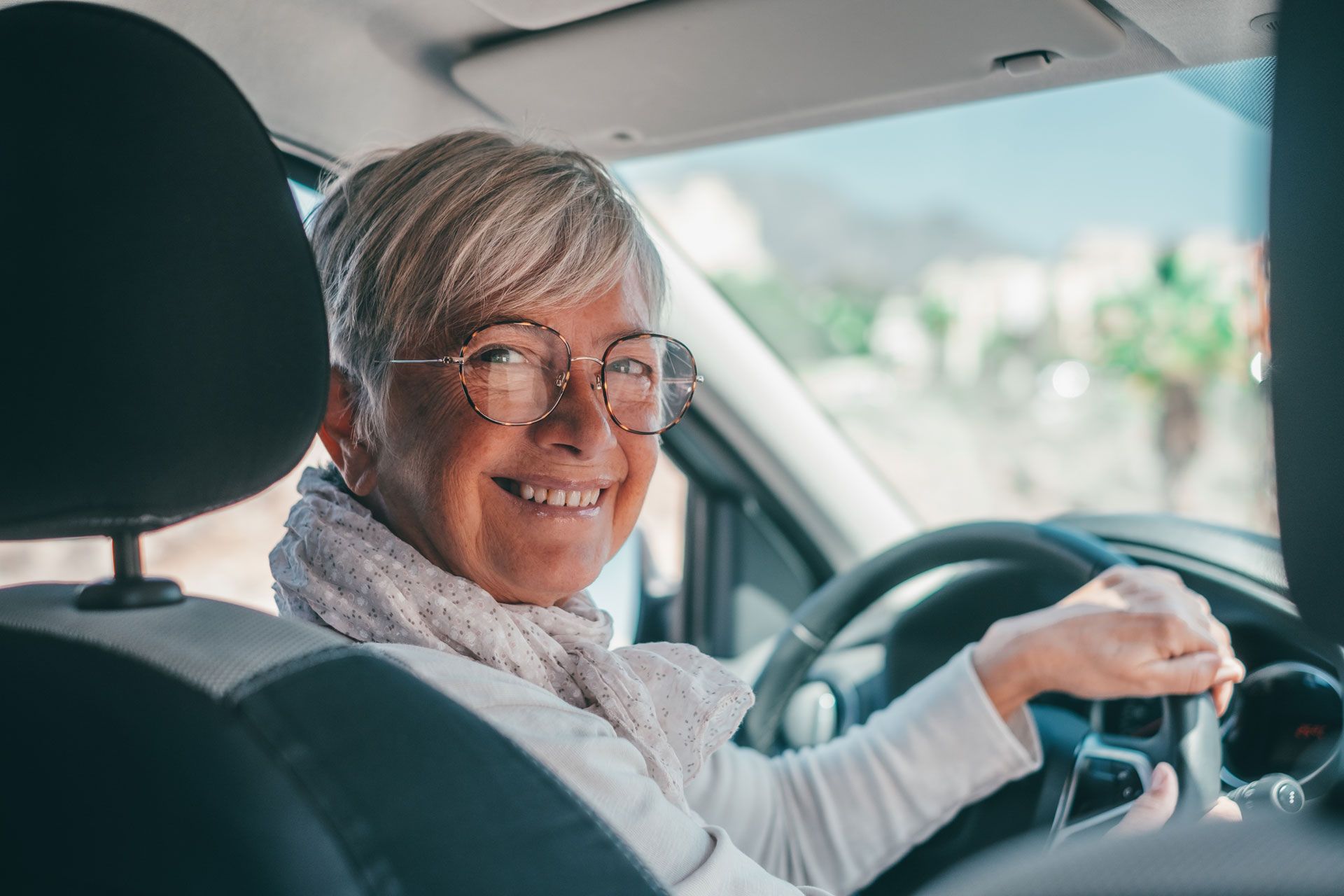 Woman smiling at the camera while driving a car; light-colored hair and glasses.