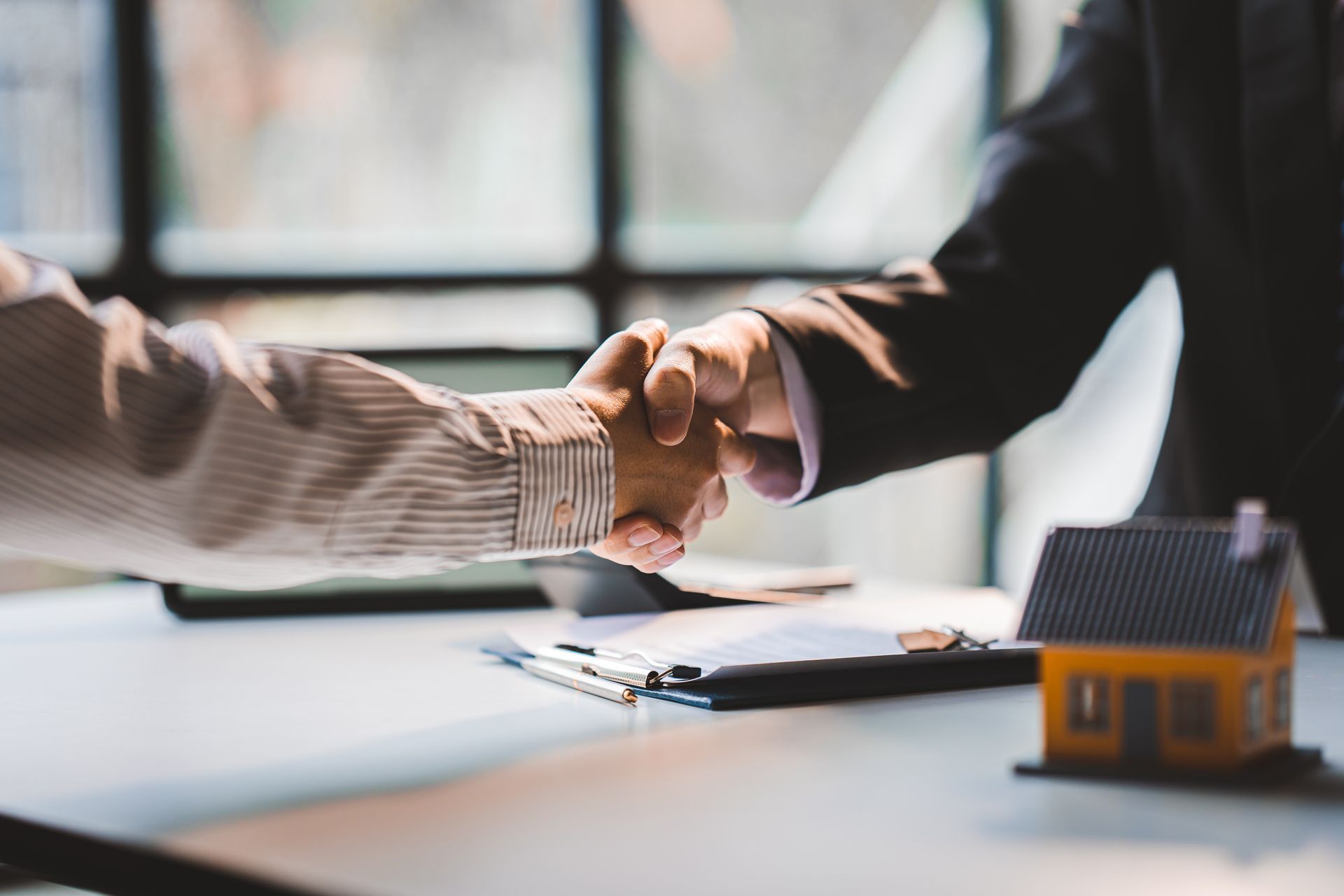 Two people shaking hands over a table with documents and a model house.