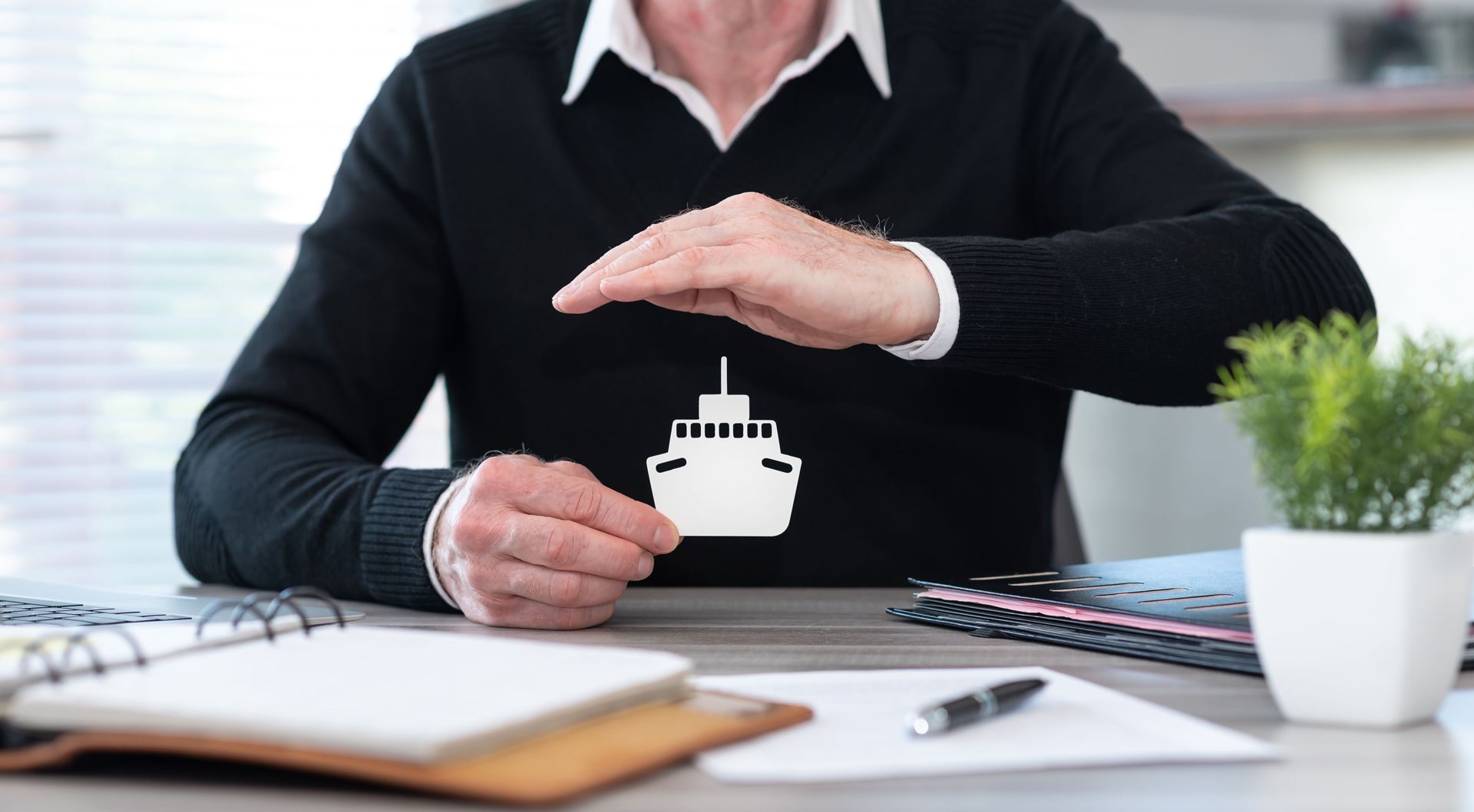 Person's hands shielding a paper ship cutout, suggesting protection, set at a desk with office supplies.