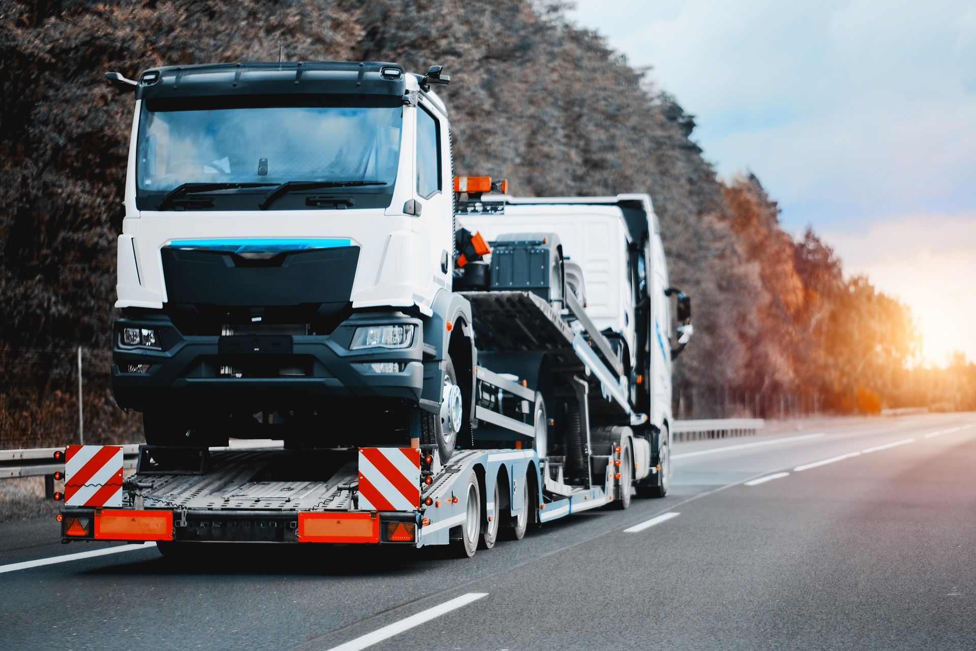 White truck carrying a smaller vehicle on a flatbed trailer, driving on a highway.