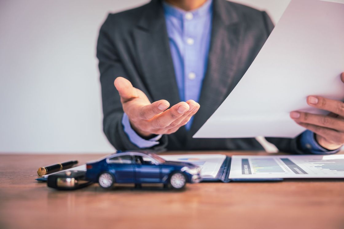 Person in suit offers papers over a toy car on a desk.
