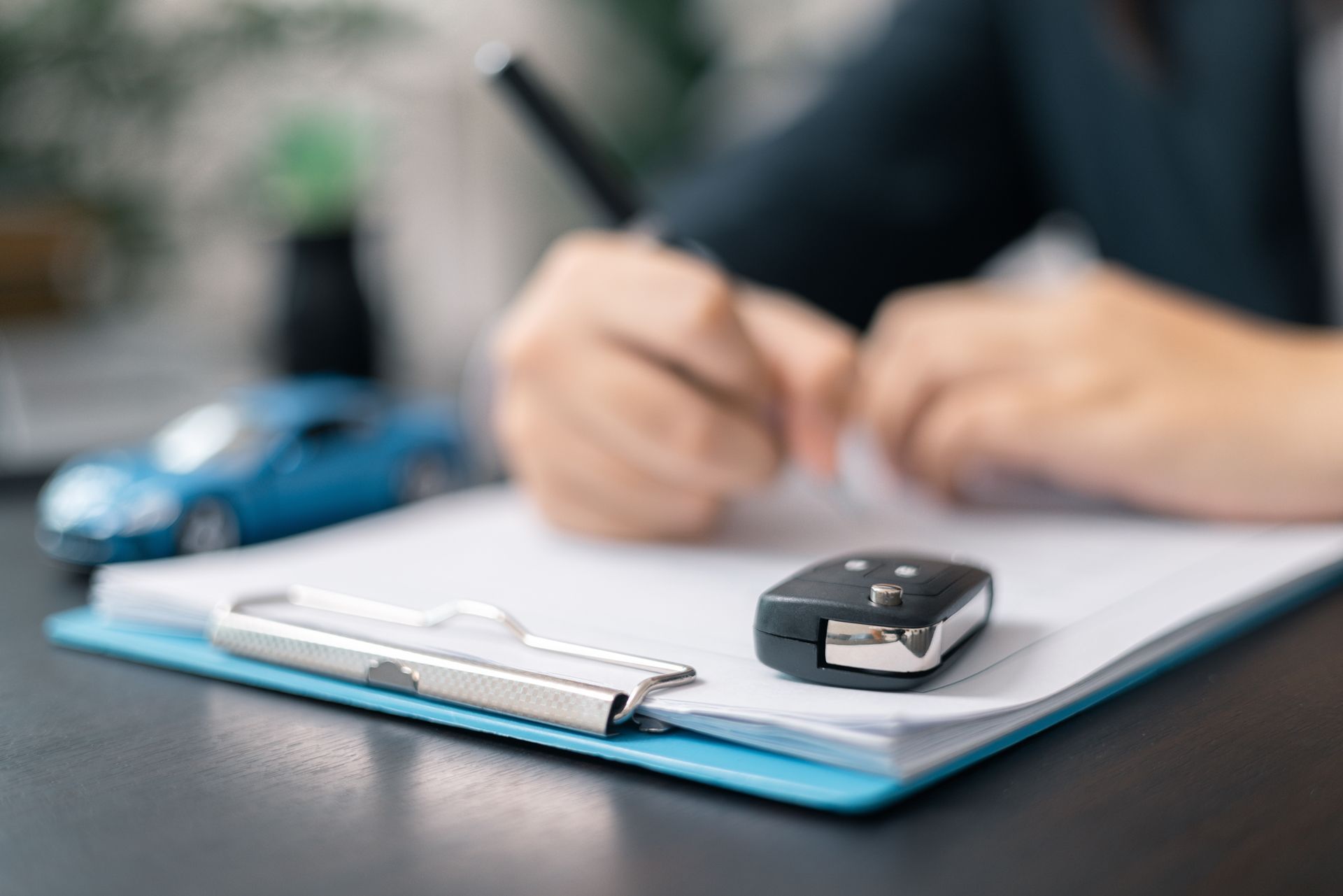 Person signing document next to car key and blue toy car on clipboard.