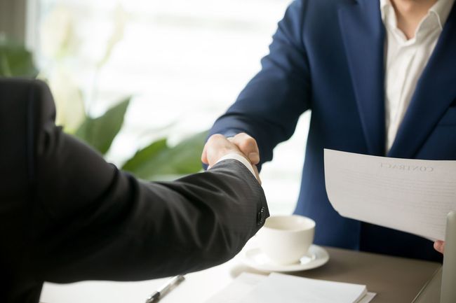 Two men in suits shaking hands at a table, documents and a coffee cup present.
