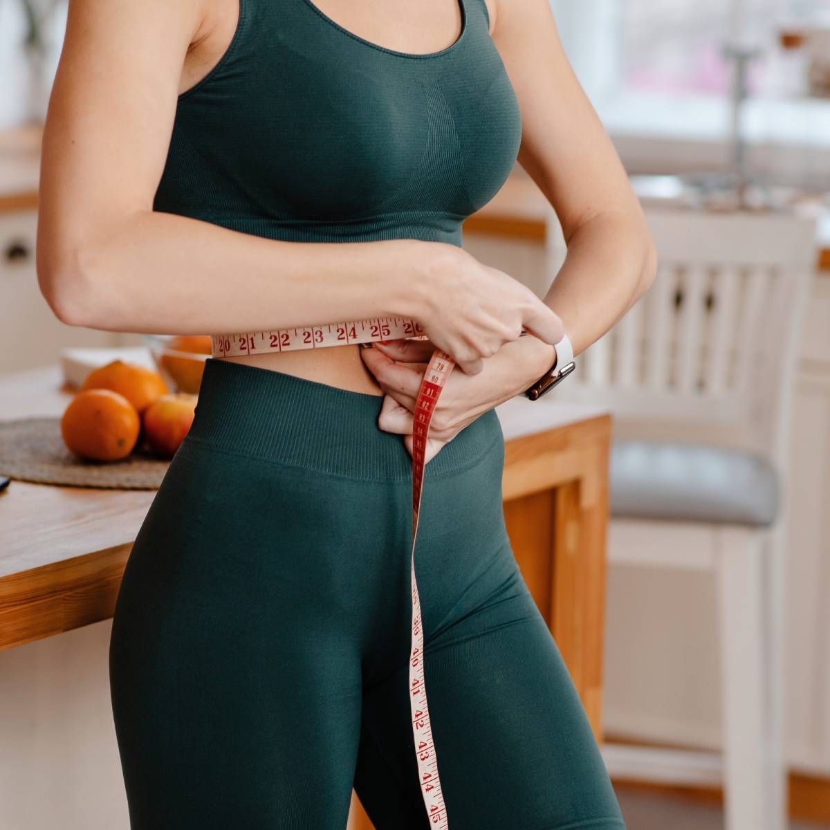 Woman in green workout clothes measures her waist with a tape measure in a kitchen.