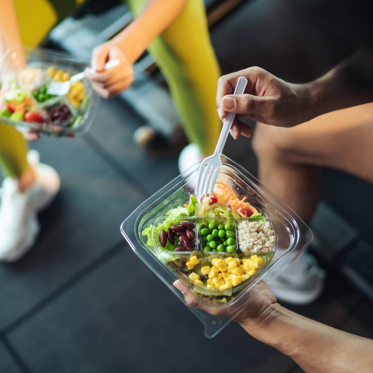 People holding salad containers, eating post-workout. Setting appears to be a gym, with colorful food.