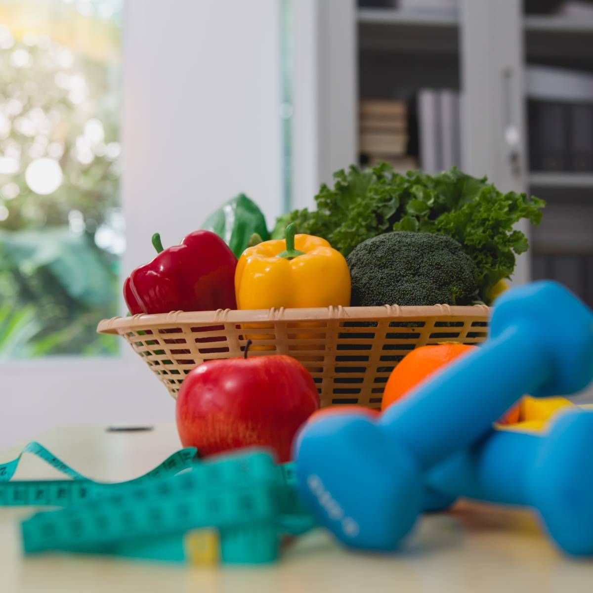 Basket of vegetables next to dumbbells, apple, orange, and a measuring tape, promoting healthy lifestyle.