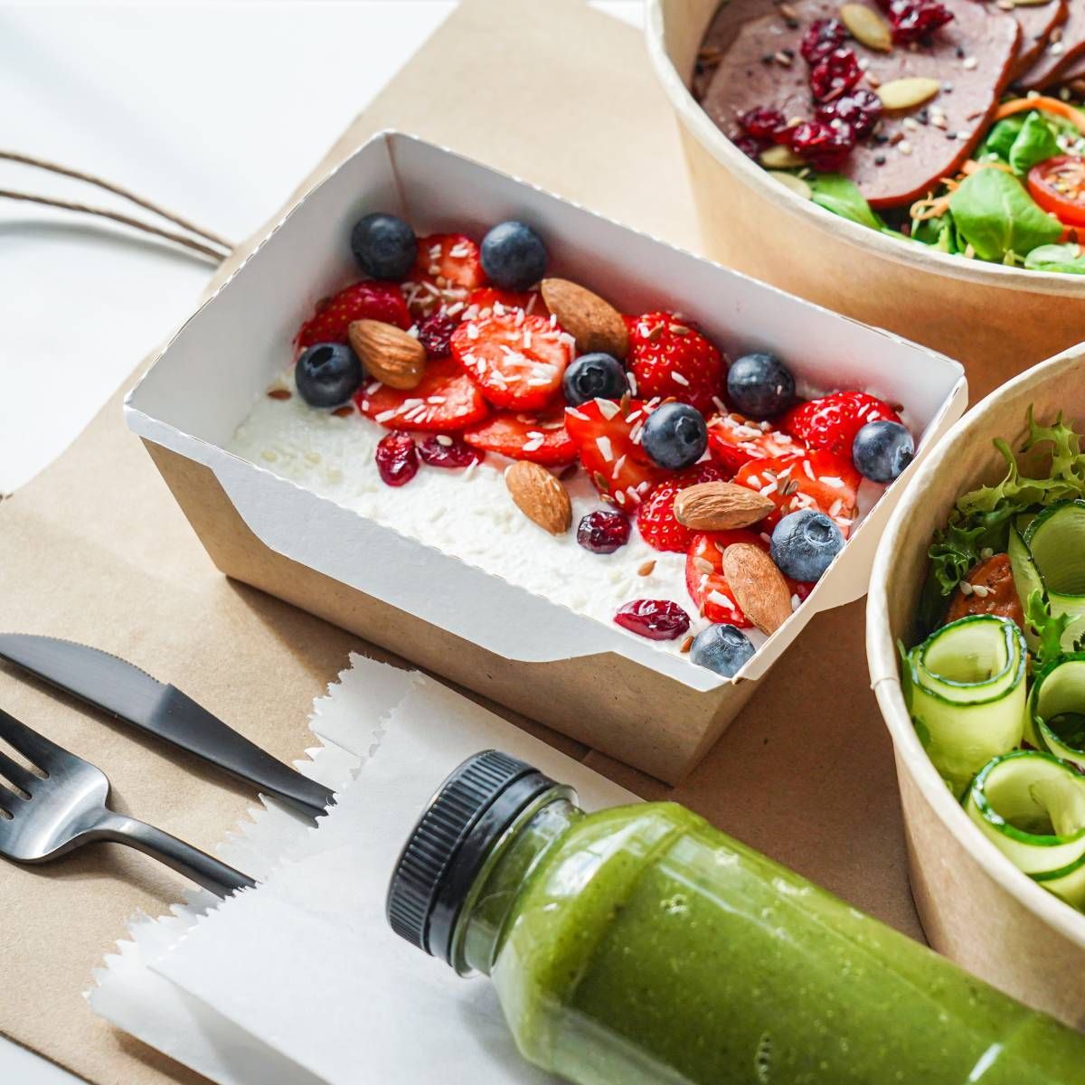 Meal prep: rice with berries and almonds, salads, green juice, cutlery on brown paper.