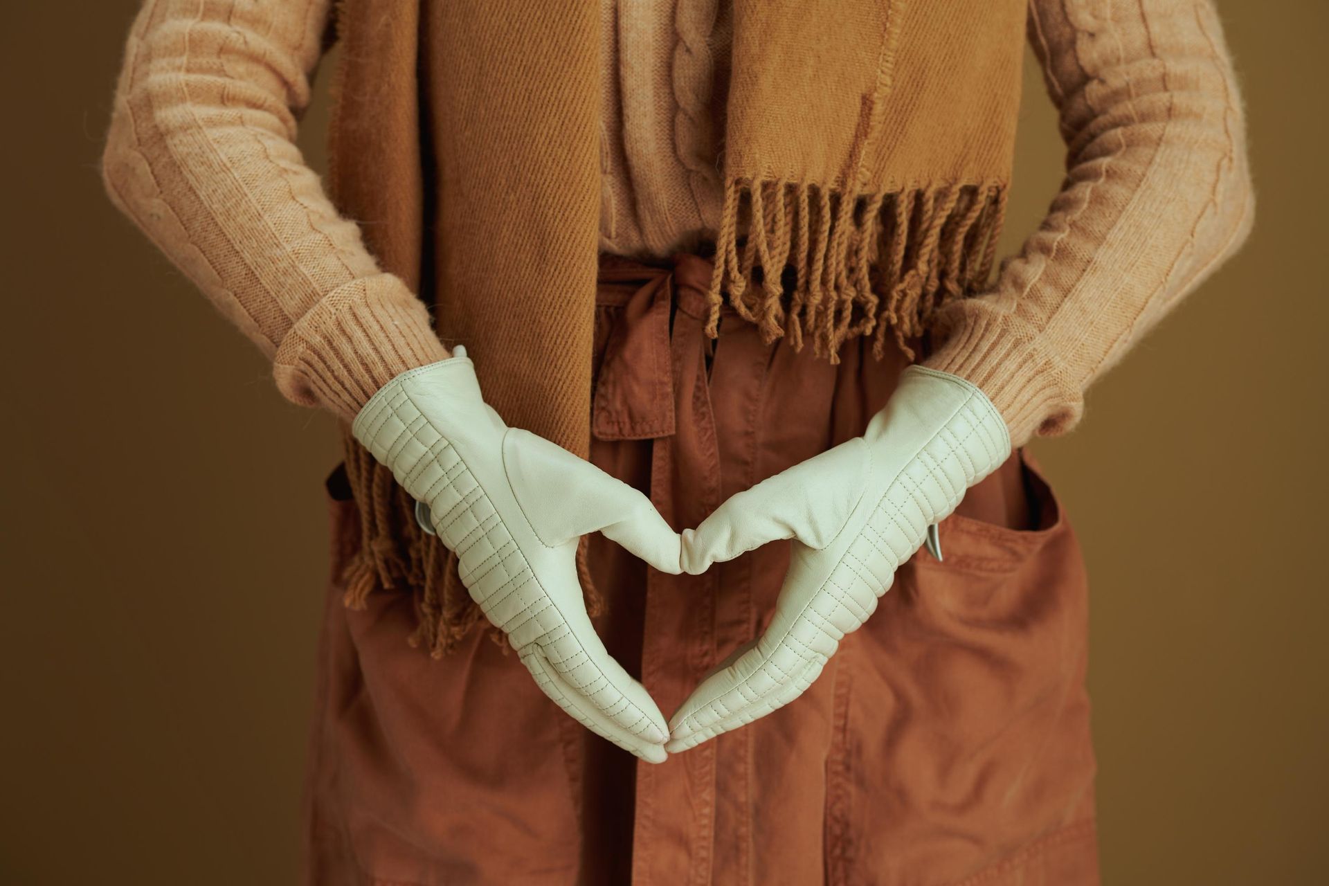 Gloved hands forming a heart shape over a brown sweater and rust-colored pants
