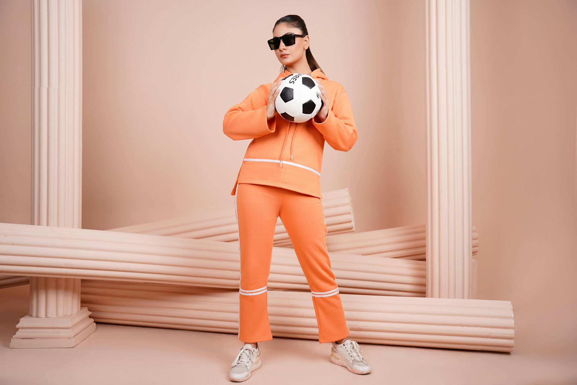 Woman in orange tracksuit holding a soccer ball, posing with sunglasses, in front of faux columns.
