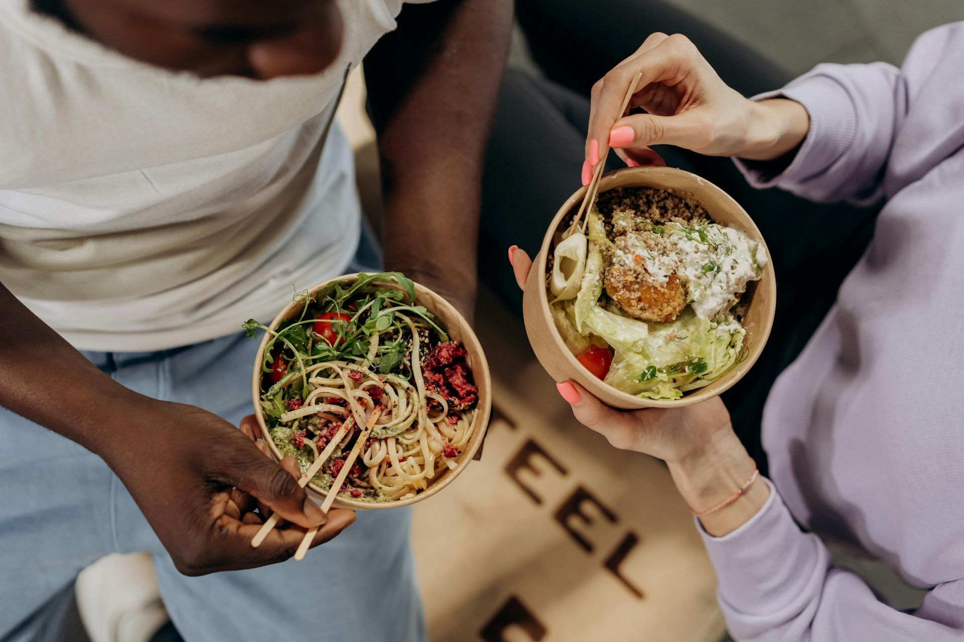 Two people holding bowls of food, one with salad, the other with a meal, seen from above.