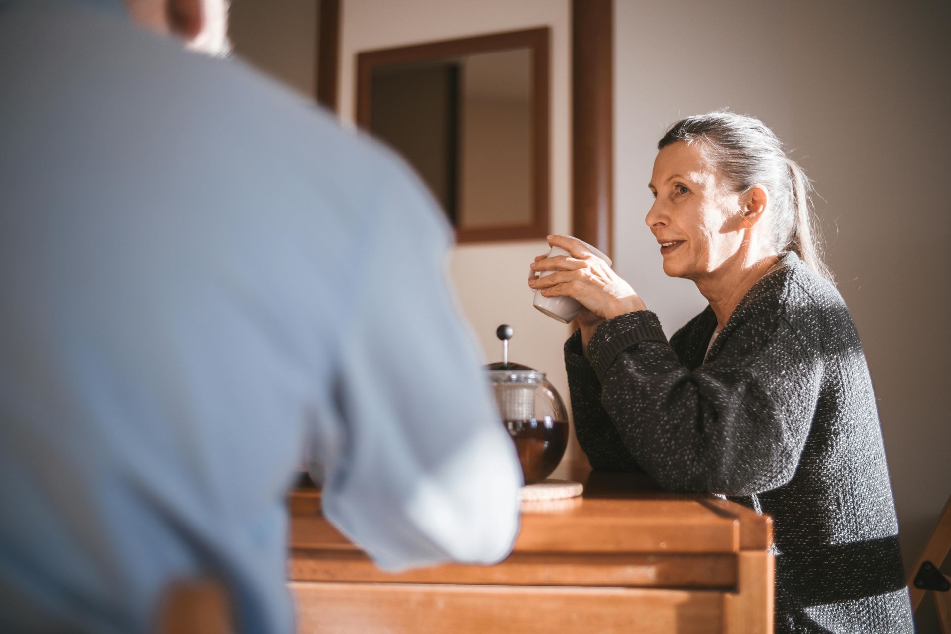 Woman with grey hair, holding a mug, talking with a person out of frame, leaning on a wooden surface.