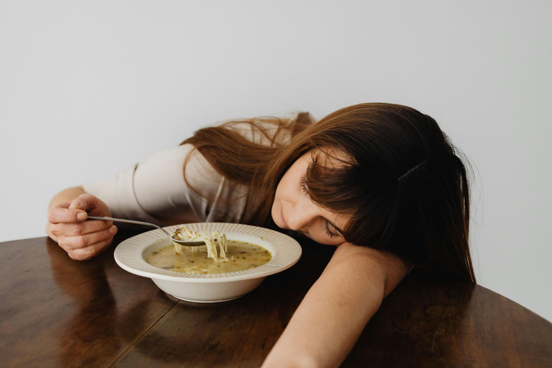 Woman slumped over a bowl of soup on a wooden table, exhausted.