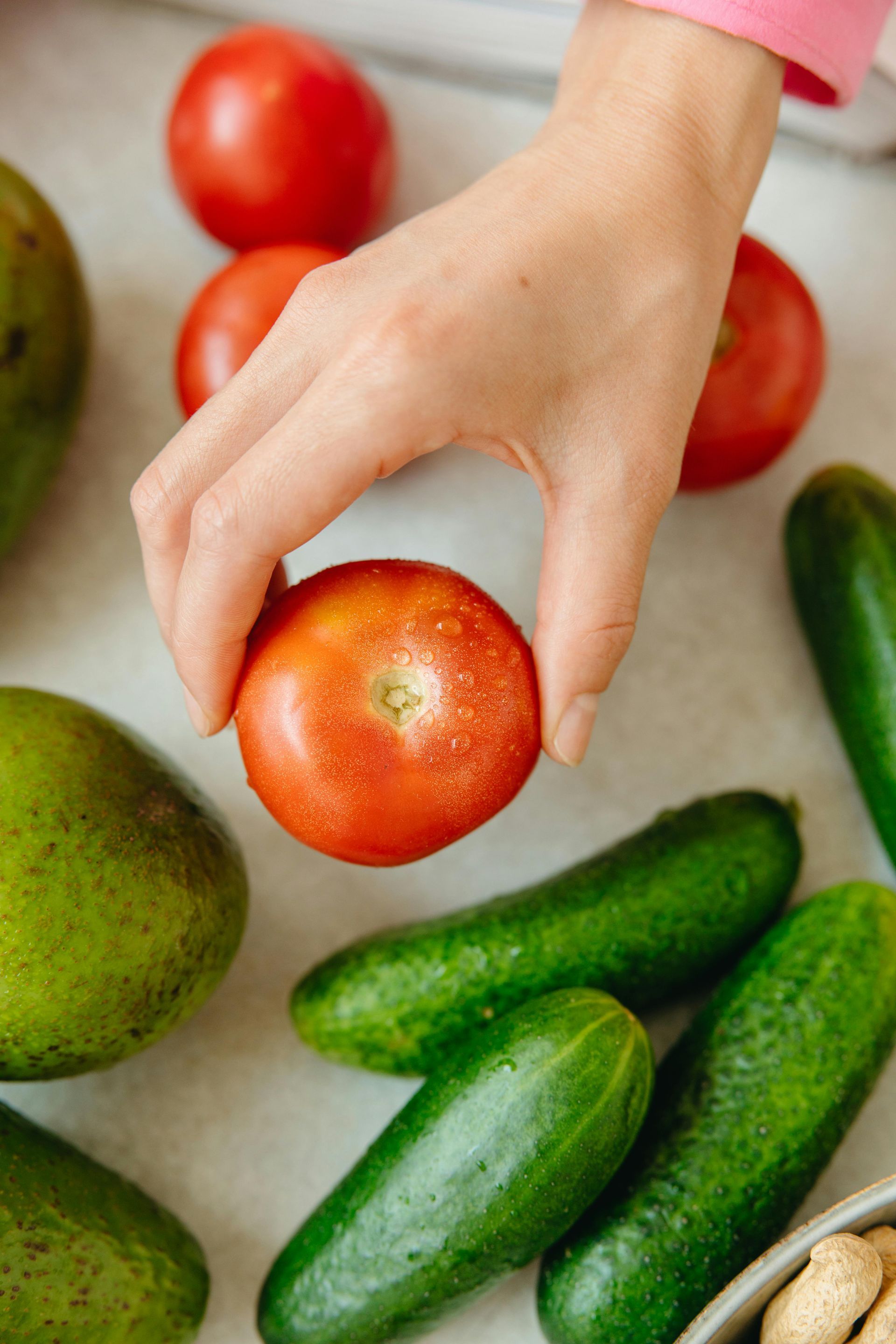 Hand holding a red tomato among cucumbers, tomatoes, and an avocado on a light surface.