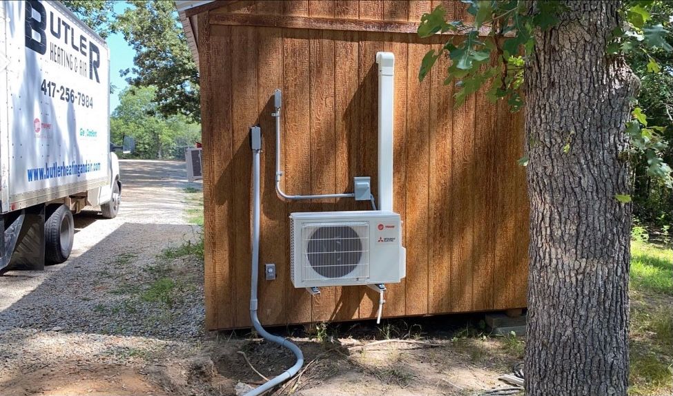 Air conditioning unit mounted on a wooden shed, with white piping and electrical conduit.