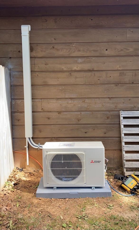 Outdoor Mitsubishi heat pump unit on a concrete pad, against a wooden wall, with conduit running upwards.