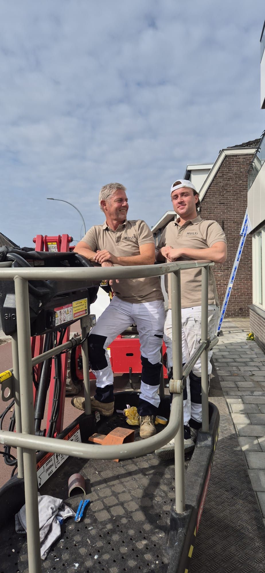 Twee schilders in beige overhemden en witte werkbroeken staan op een hoogwerker buiten een bakstenen gebouw.