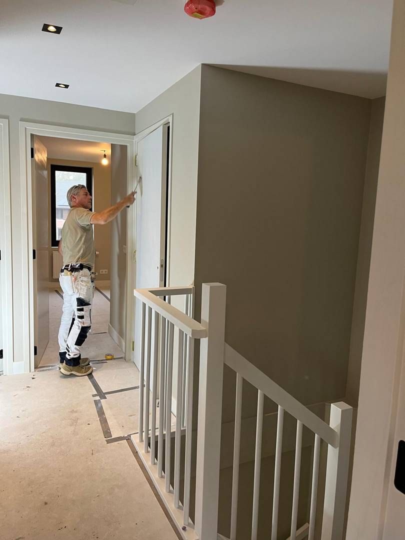 Een werknemer in een werkbroek en een T-shirt plakt tape op een witte muur in een gang naast een trappenhuis.
