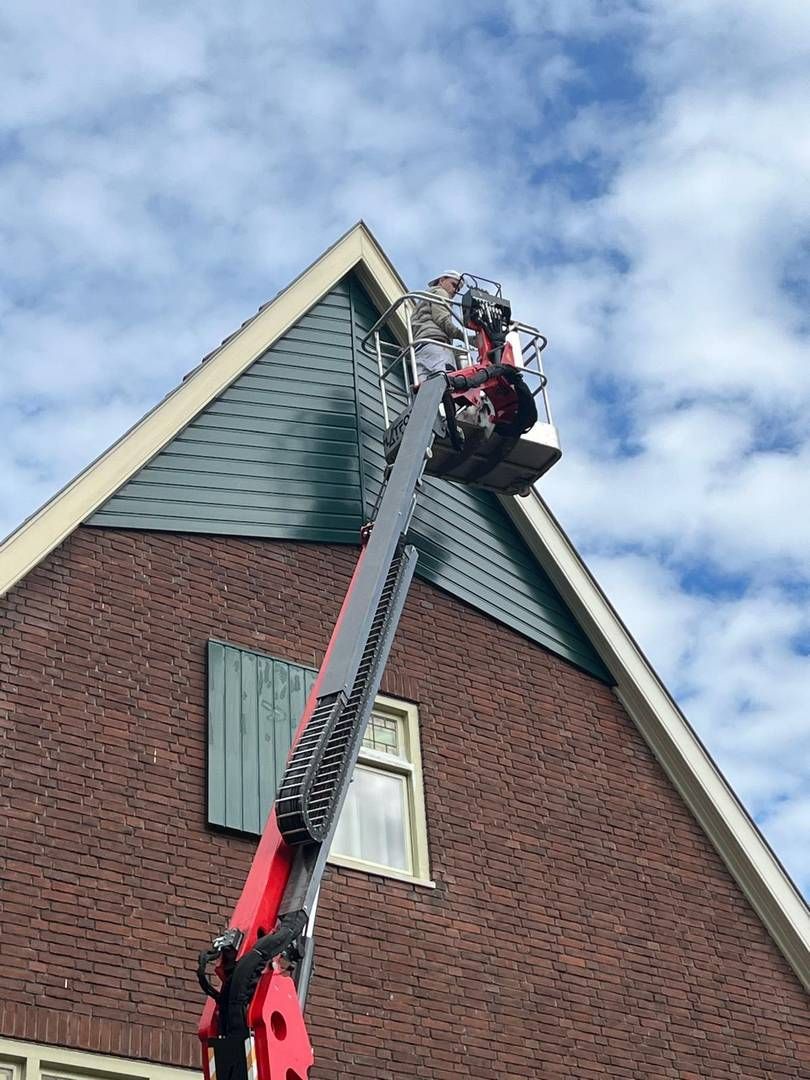 Een persoon in een rode werkjas staat in de cabine van een hoogwerker en reikt naar de top van een bakstenen gebouw.