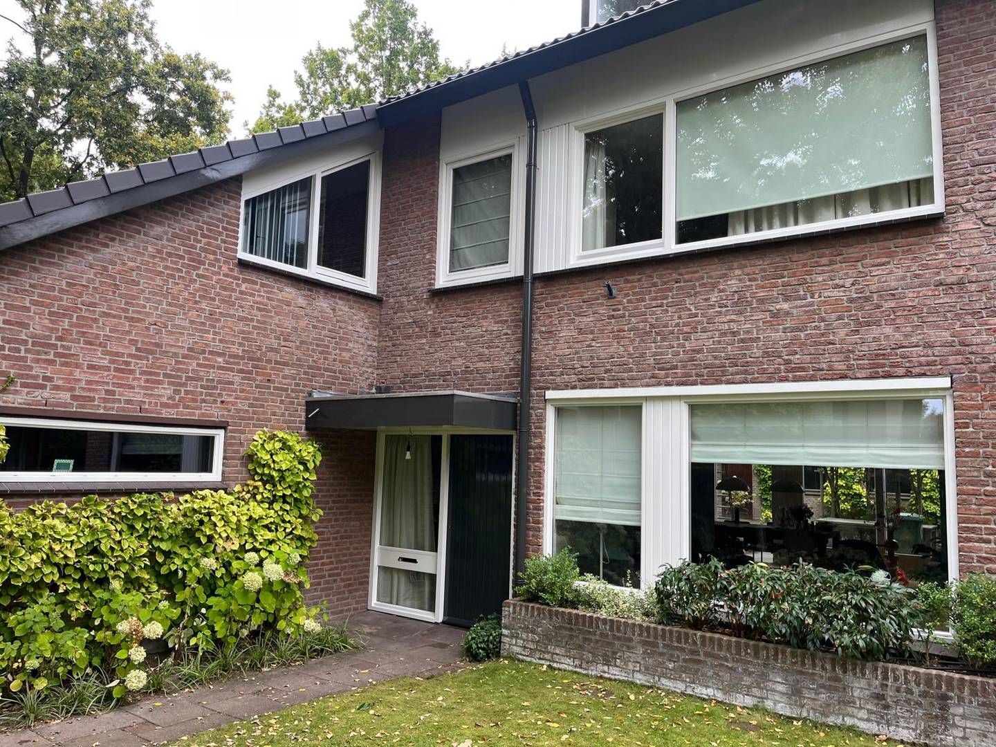 Een bakstenen huis met twee verdiepingen, een plat dak, grote ramen, een kleine voortuin en een overdekte entree.
