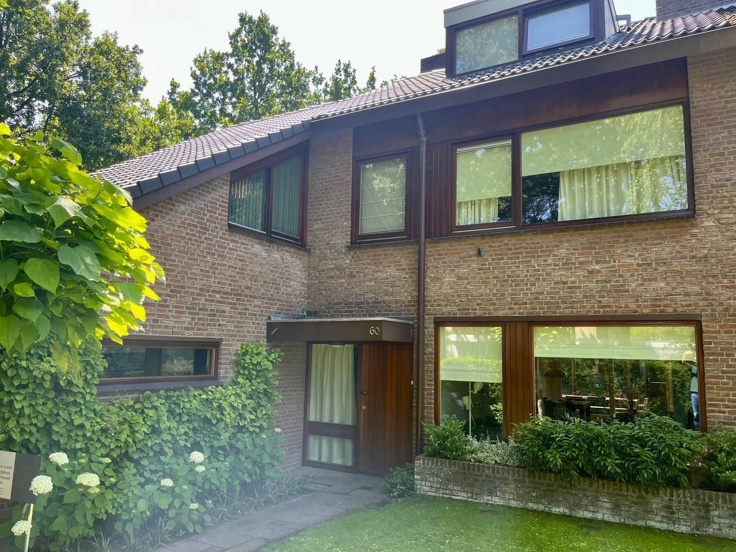 Een twee verdiepingen tellend huis van bruine baksteen met een zadeldak, grote ramen en een aangelegde tuin aan de voorzijde.