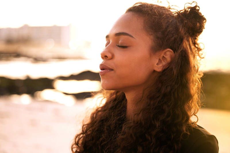 A woman is standing in front of a body of water with her eyes closed.