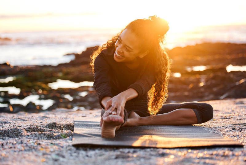 A woman is sitting on a yoga mat on the beach stretching her legs.