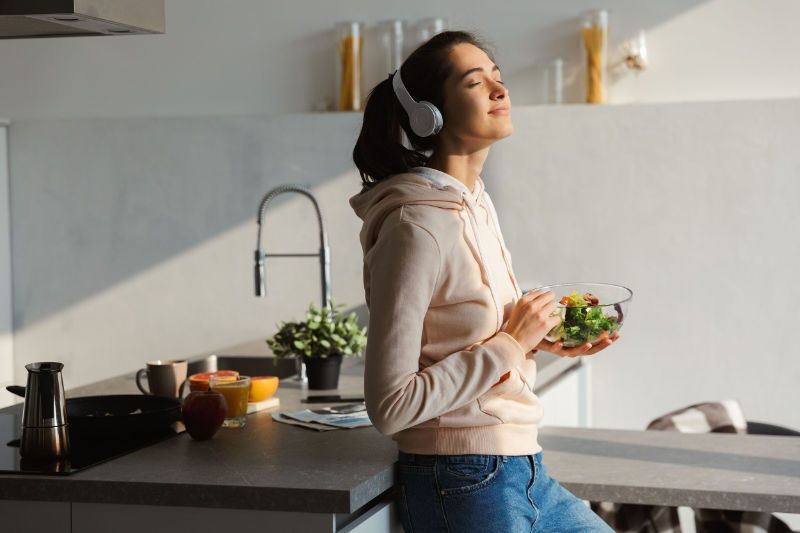 A woman wearing headphones is standing in a kitchen holding a bowl of salad.