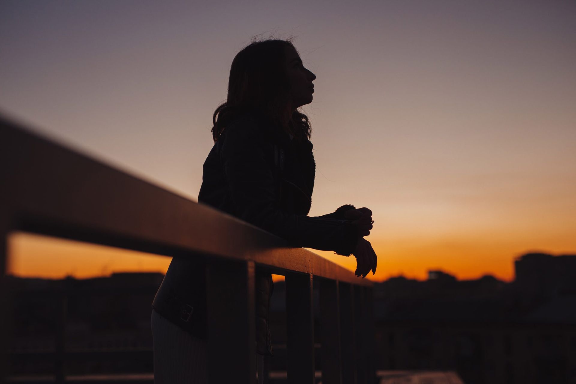 A silhouette of a woman standing on a balcony at sunset.