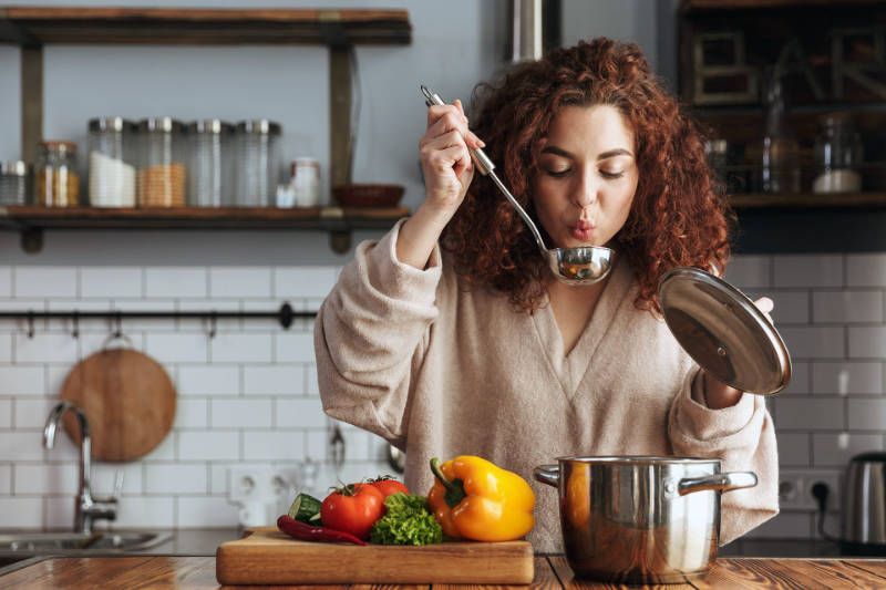 A woman is eating soup from a ladle in a kitchen.