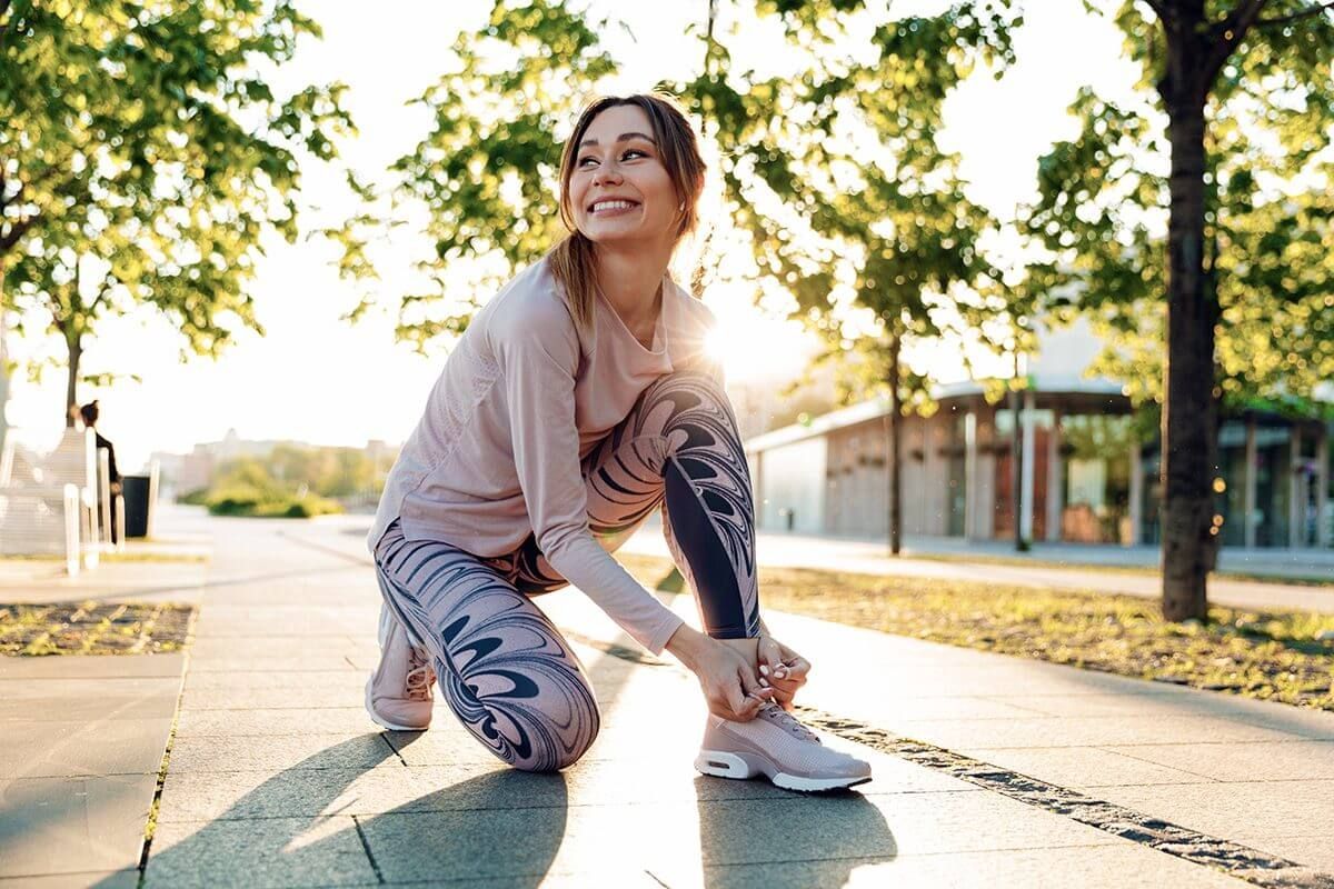 A woman is kneeling down on the sidewalk tying her running shoes.