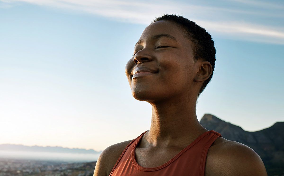 A woman is standing in front of a mountain with her eyes closed smiling.