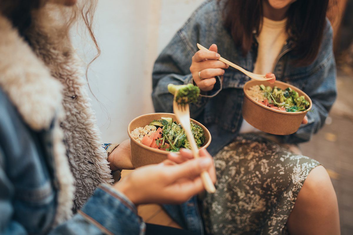 Two women are sitting at a table eating food from bowls with chopsticks.