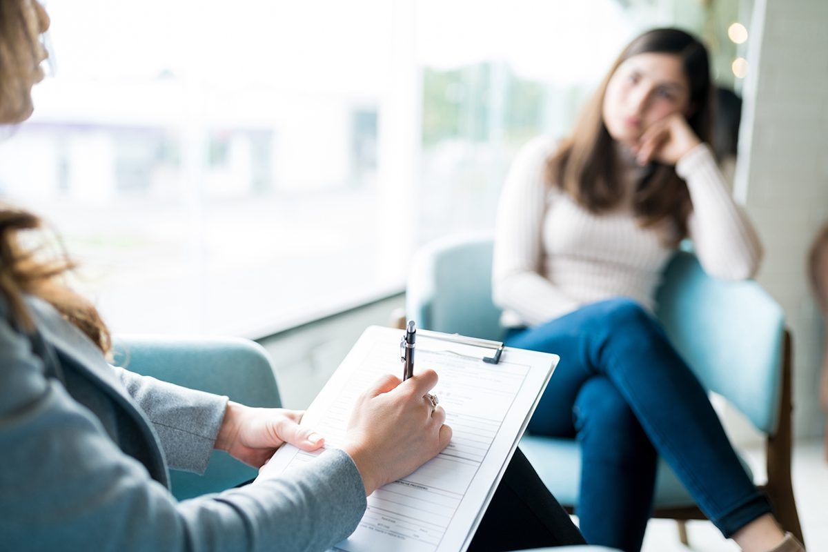 A woman is sitting in a chair talking to another woman while holding a clipboard.