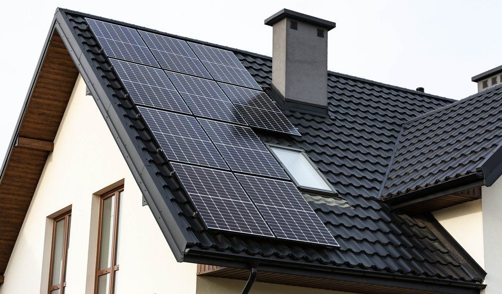 Solar panels on a black-tiled roof of a white house, next to a chimney and skylight.